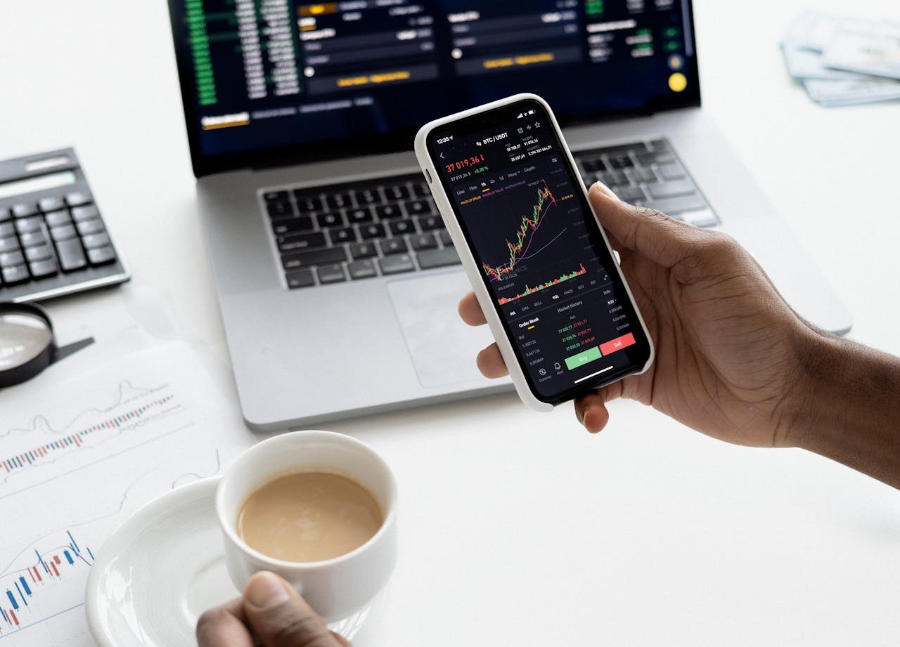 A person trades stocks on a smartphone with a laptop, coffee cup, calculator, and documents in view.